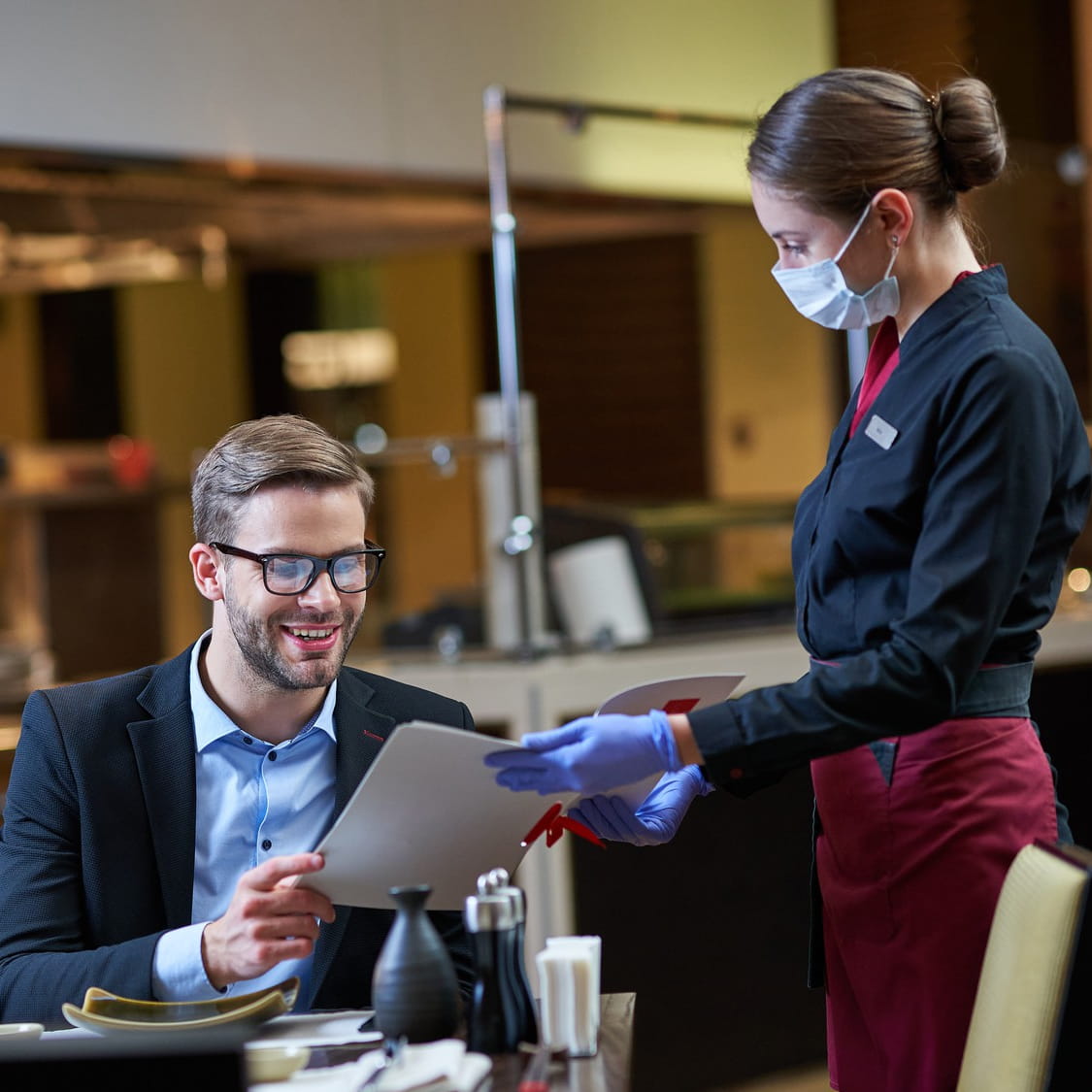 Server wearing a mask handing a menu to a seated restaurant guest